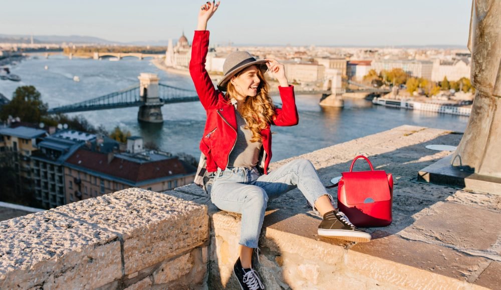 Adorable young female model with light-brown hair expressing happy emotions, traveling in Europe. Blissful girl in trendy red jacket and hat enjoying photoshoot on river background in city.